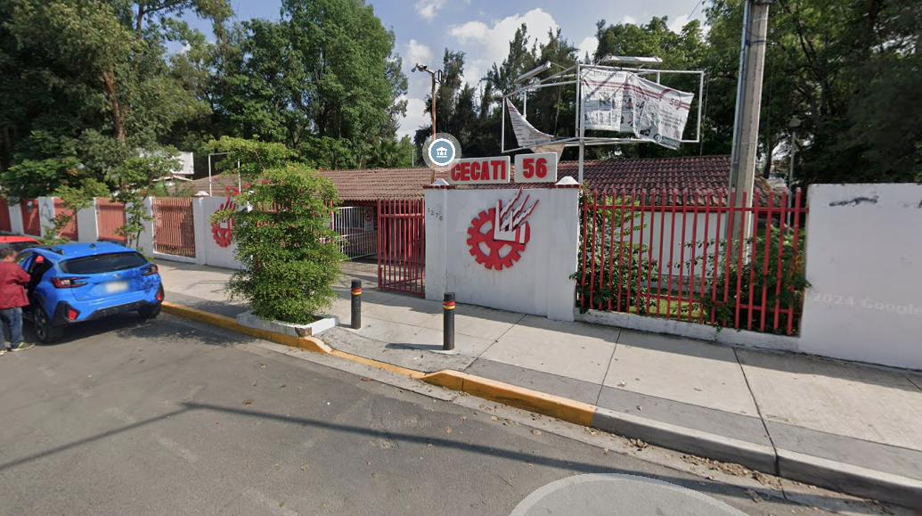 Fotografía en la que se muestra la entrada del plantel, conformada por una reja de color rojo que da acceso a las instalaciones. A la derecha de la reja hay un muro blanco en el que está el logotipo del plantel. Arriba del muro hay un cartel que dice CECATI 56. El plantel está rodeado por rejas rojas intercaladas con muros blancos.